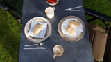 Outdoor setting with plates of food on a tablecloth, surrounded by grass.