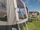 Person sitting at a table with a dog next to a caravan with an extended awning on a grassy area.