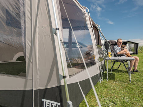 Person sitting at a table with a dog next to a caravan with an extended awning on a grassy area.